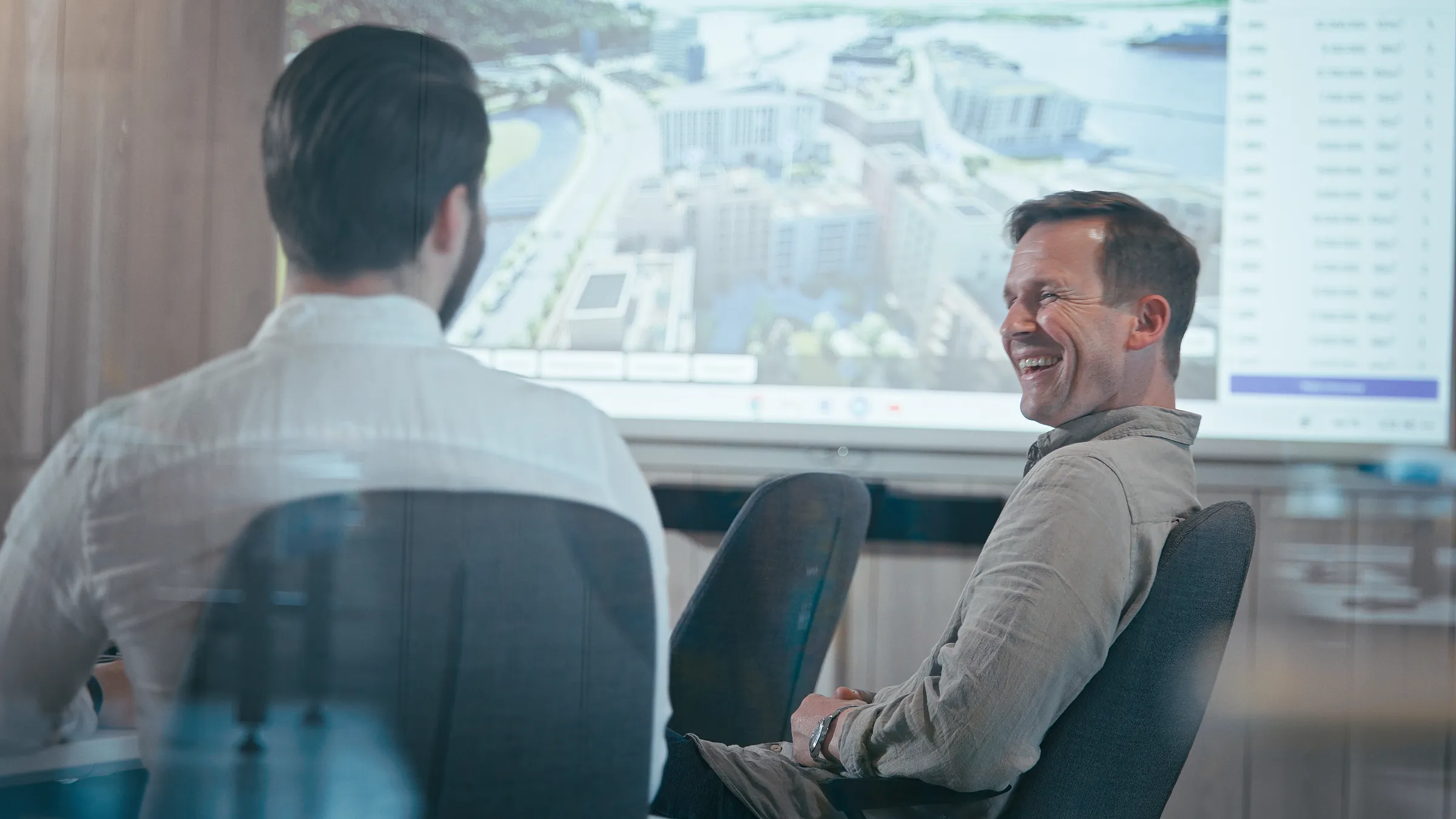 Two colleagues laughing in a meeting room, a housing project on the screen behind them
